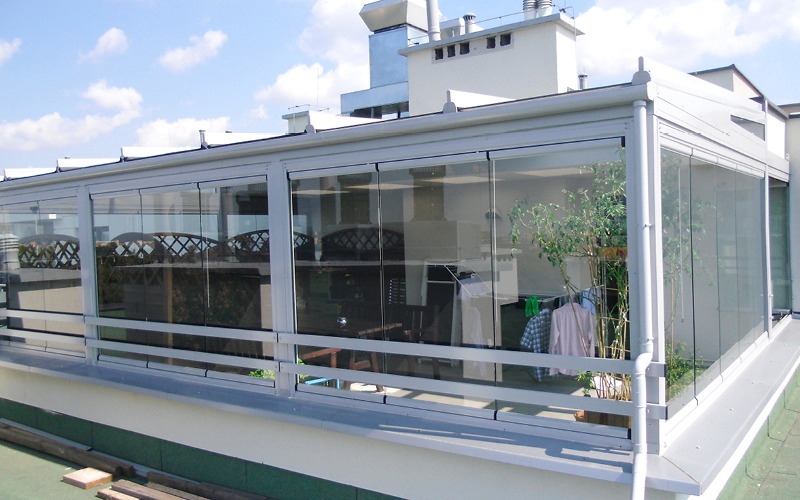 Przeszklona zabudowa balkonu z przesuwnymi panelami, widok z zewnątrz, w tle budynki mieszkalne i niebo, na balkonie suszarka z praniem i roślina w donicy.