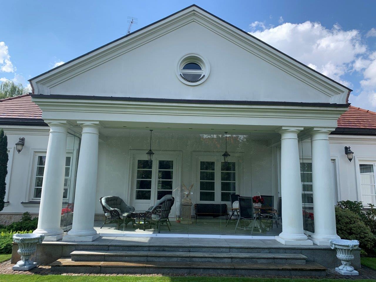 Elegancki taras z kolumnami i przeszklonymi balustradami, meble ogrodowe, widok na budynek z oknem w kształcie okręgu