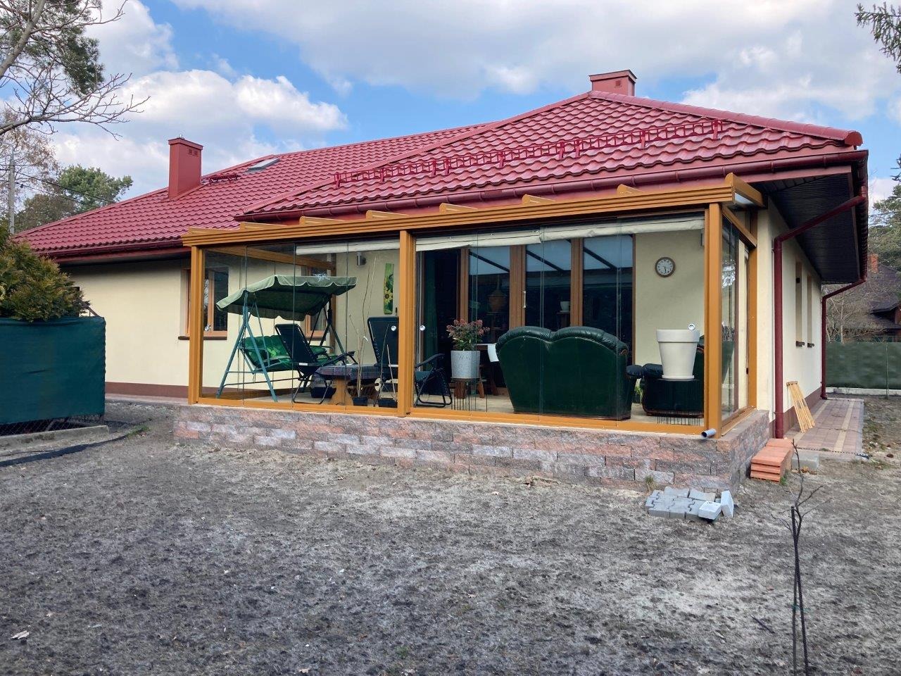 Przeszklona weranda z drewnianą konstrukcją, przylegająca do domu z czerwoną dachówką, wyposażona w meble ogrodowe i rośliny doniczkowe, widok z zewnątrz.