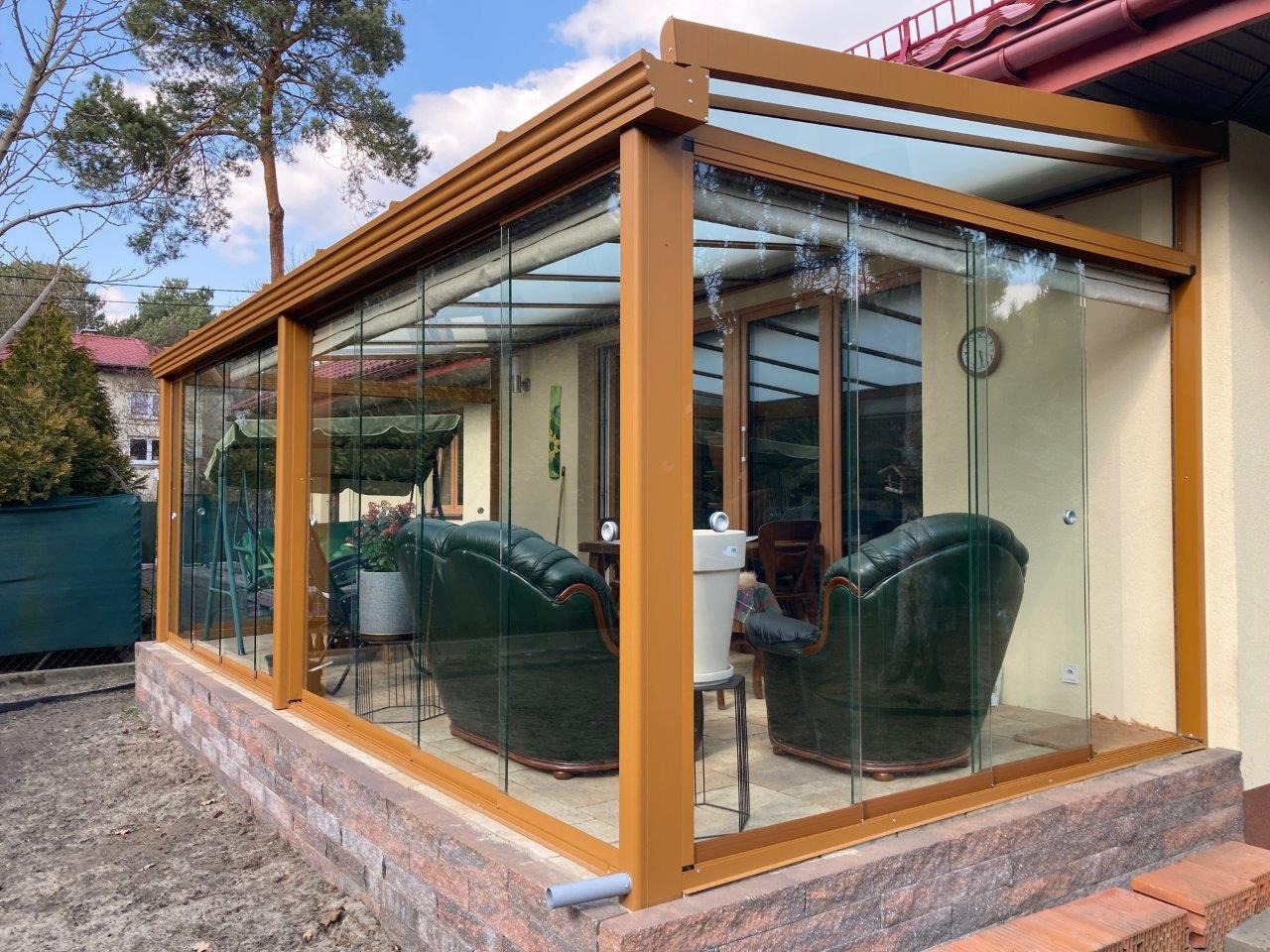 Zabudowany taras z brązową ramą i szklanymi ścianami, widoczne zielone fotele i ceglana podmurówka, częściowo wykończone otoczenie.