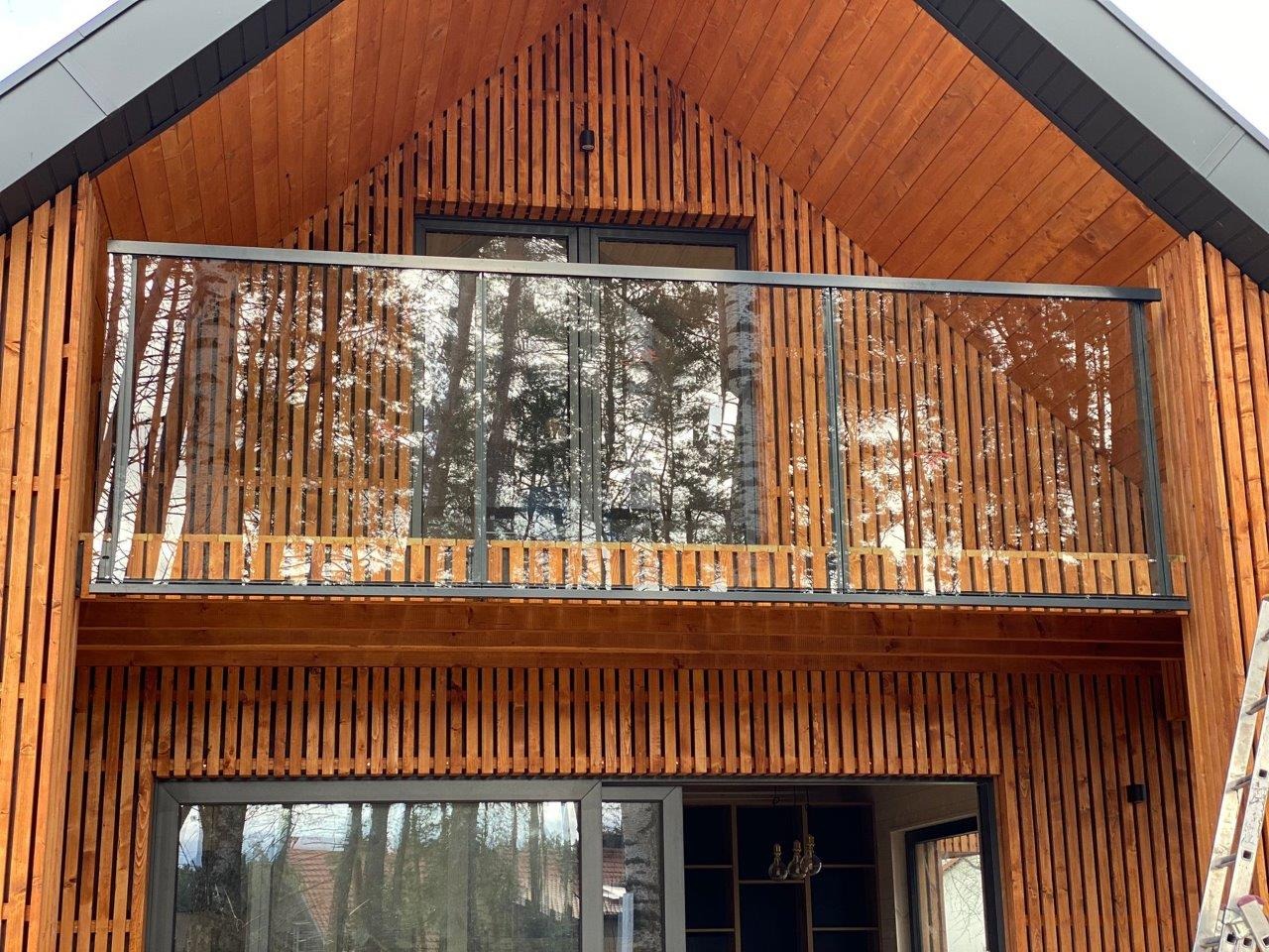 Nowoczesny dom z drewnianą elewacją i balkonem zabezpieczonym szklaną balustradą z czarnymi ramami, widok z dołu na tle drzew.