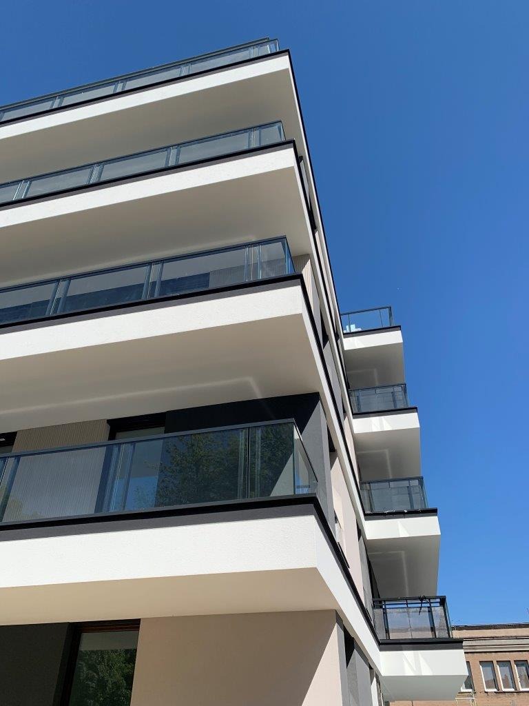 Nowoczesny budynek wielorodzinny z balkonami zabezpieczonymi szklanymi balustradami, ujęcie z dołu w słoneczny dzień, widoczne odbicie drzew w szkle.