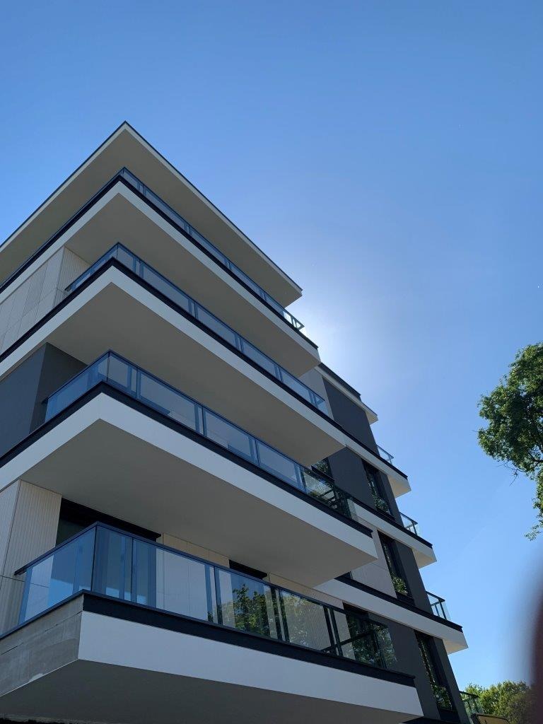 Nowoczesny budynek wielorodzinny z balkonami zabezpieczonymi szklanymi balustradami, widok z dołu na błękitne niebo.