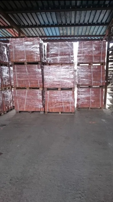 Sztaple cegieł ceramicznych na paletach, zabezpieczone folią, składowane w magazynie budowlanym z metalową konstrukcją dachu.