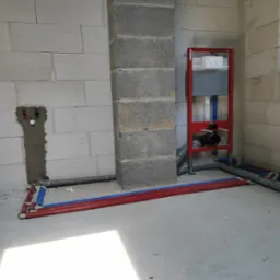 Instalacja podtynkowa WC ze stelażem w czerwonej ramie, widoczne rury doprowadzające wodę w izolacji koloru czerwonego i niebieskiego, ściany z białego betonu komórkowego.