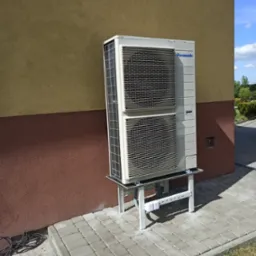 Zewnętrzna jednostka pompy ciepła Panasonic zamontowana na metalowym stelażu przy dwukolorowej elewacji budynku i chodniku z kostki brukowej.