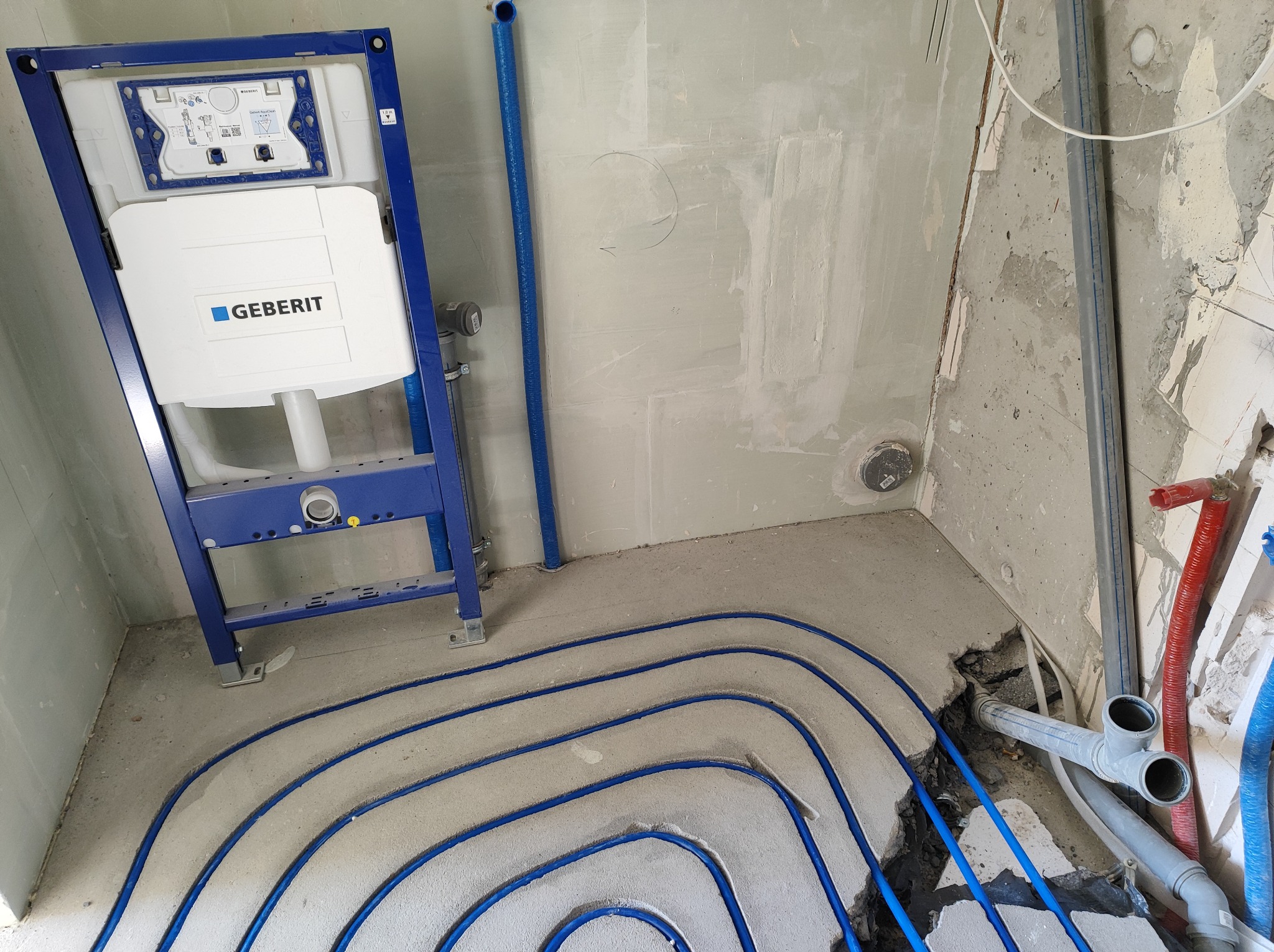 Stelaż podtynkowy Geberit zamontowany w surowym wnętrzu, z widocznym ogrzewaniem podłogowym w postaci niebieskich rurek zatopionych w wylewce, oraz fragmenty instalacji kanalizacyjnej i wodnej.