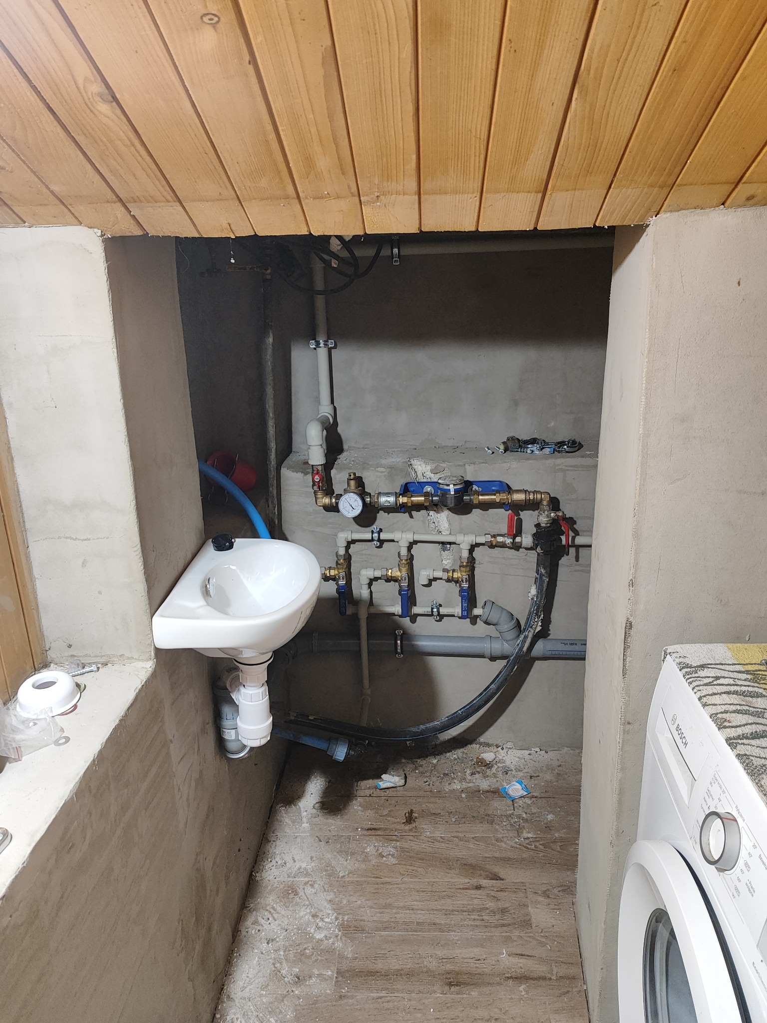 Narożna umywalka zamontowana w surowym wnętrzu z widoczną instalacją hydrauliczną, rurami i zaworami; obok pralka.