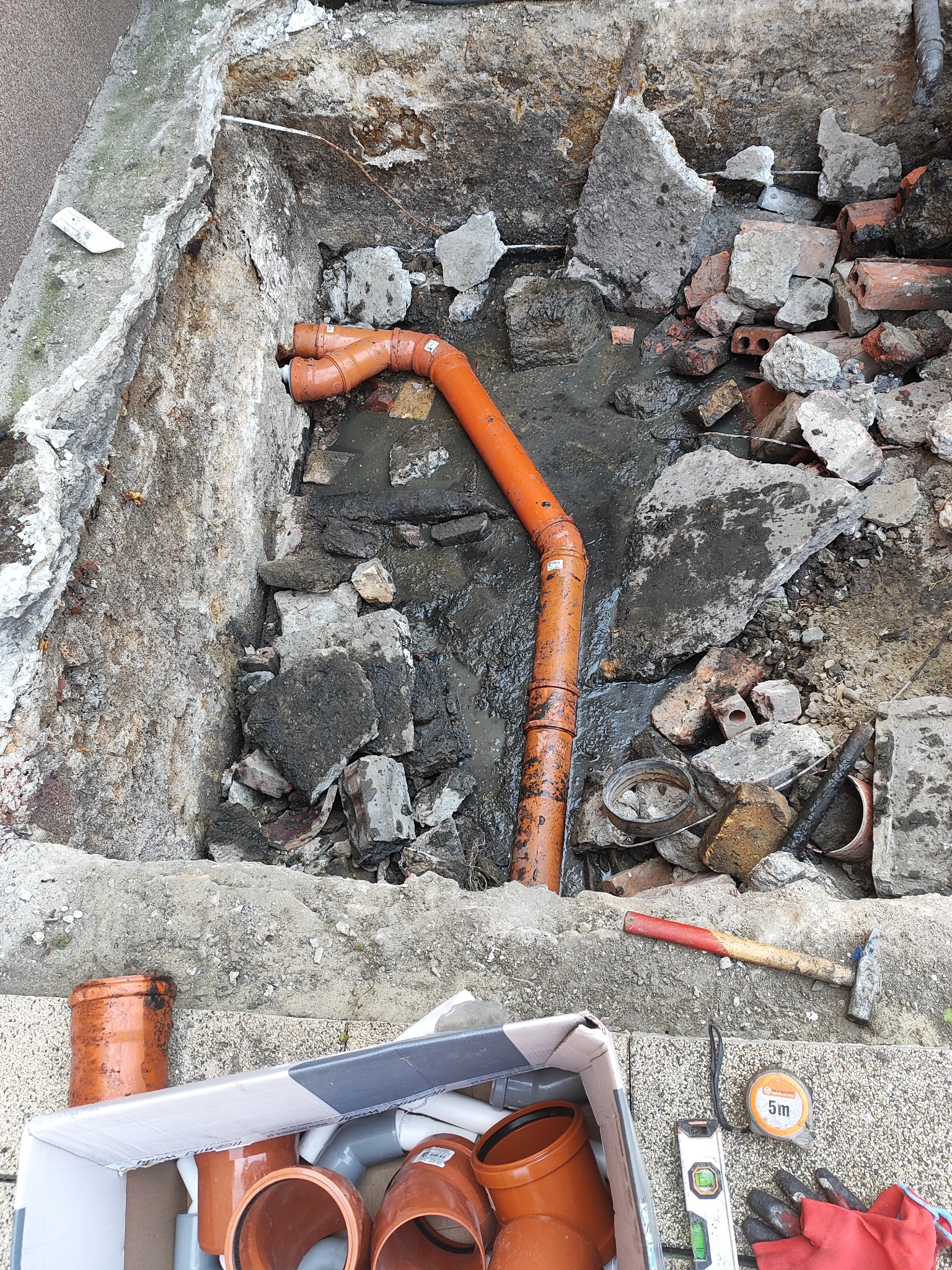 Wykop z pomarańczowymi rurami kanalizacyjnymi, gruzem i narzędziami: młotek, poziomica, miarka zwijana, rękawice robocze, karton z kształtkami.