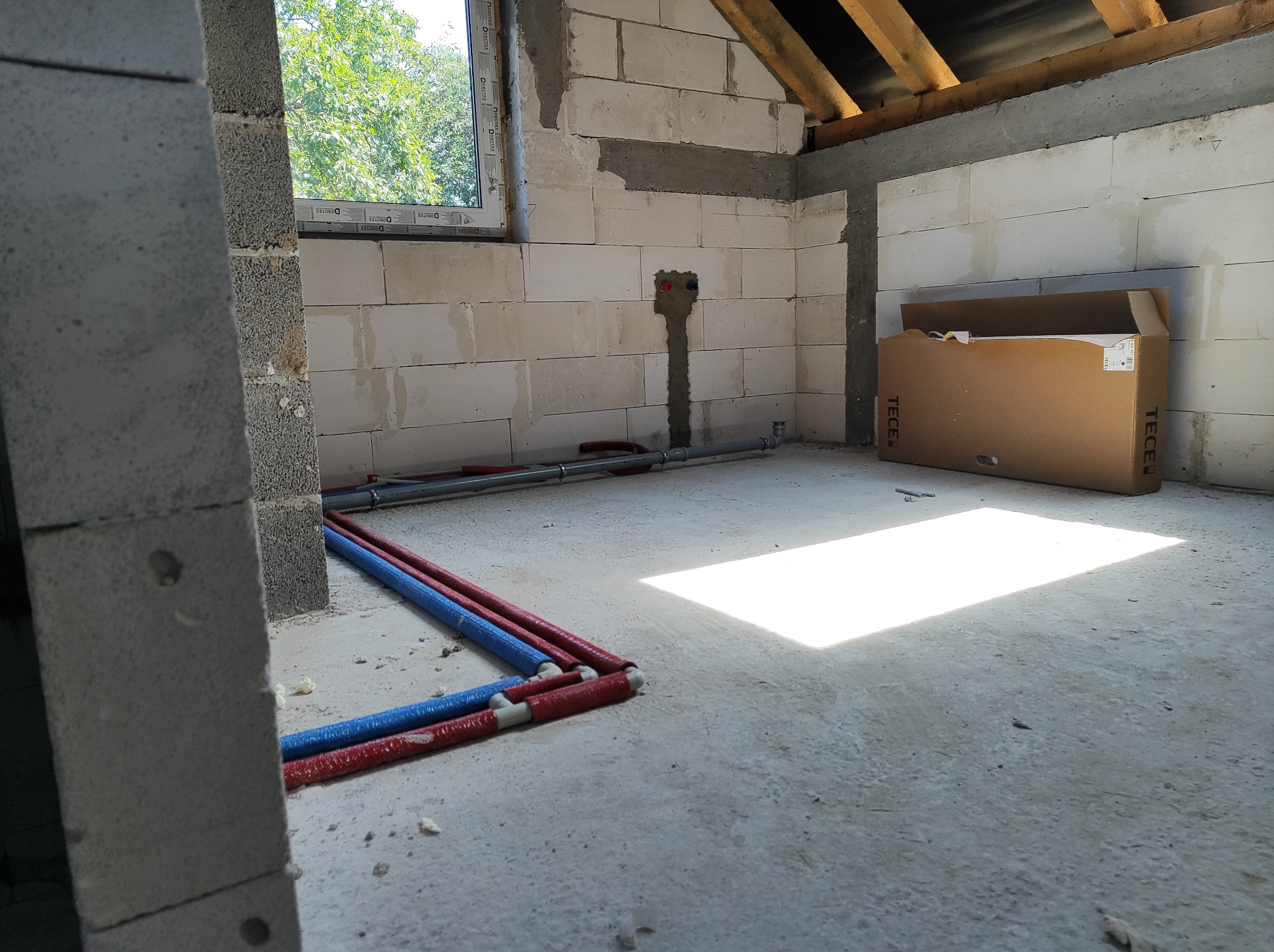 Surowy stan: Instalacja hydrauliczna z czerwono-niebieskich rur w nieotynkowanym pomieszczeniu z oknem, widoczne przyłącza w ścianie i karton TECE