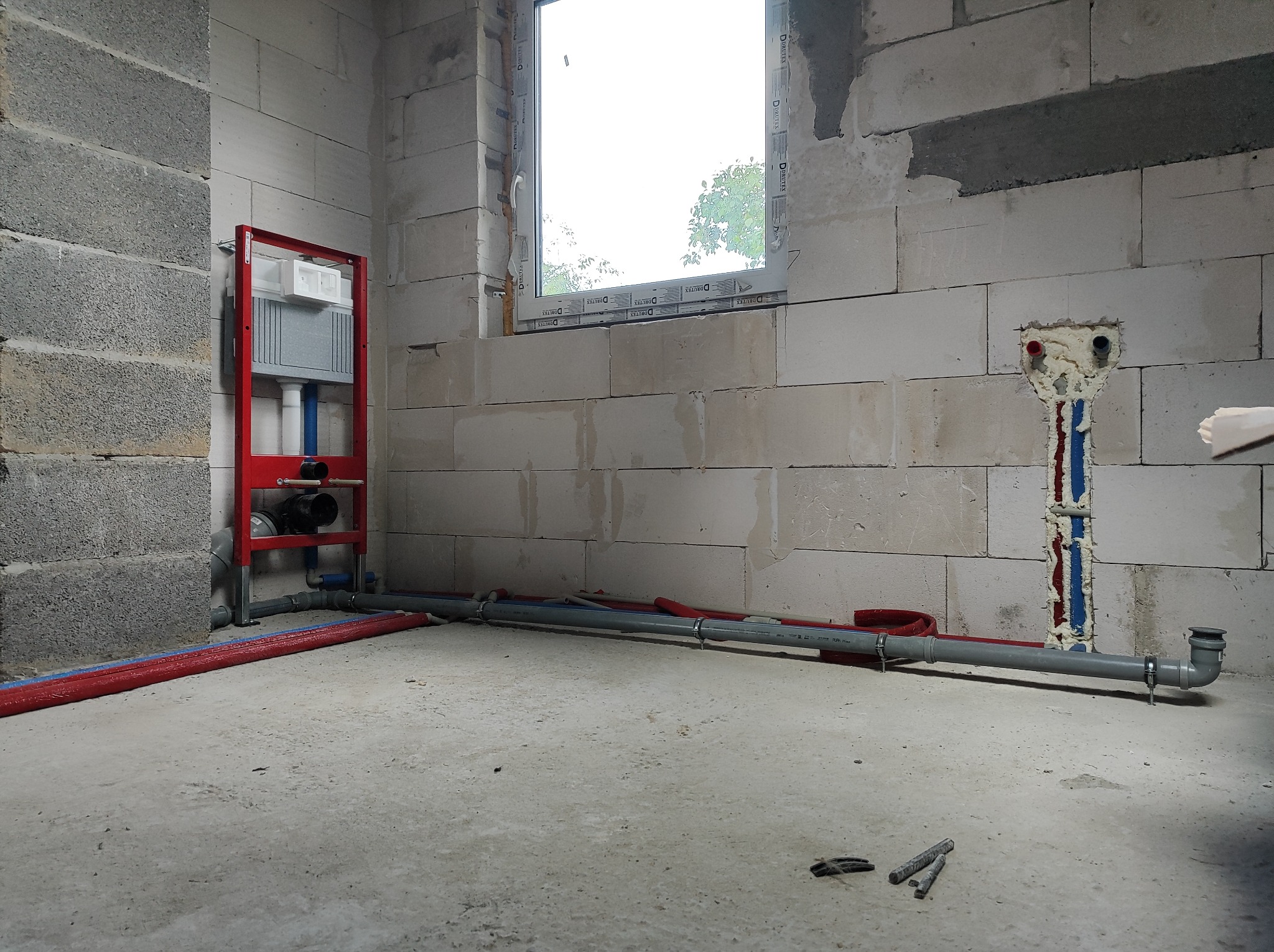 Surowy stan łazienki: widoczny stelaż podtynkowy WC w czerwonej ramie, rury instalacyjne w podłodze i ścianie, oraz otwór w ścianie z uszczelnionymi rurami. Ściany z bloczków betonowych, okno w tle.