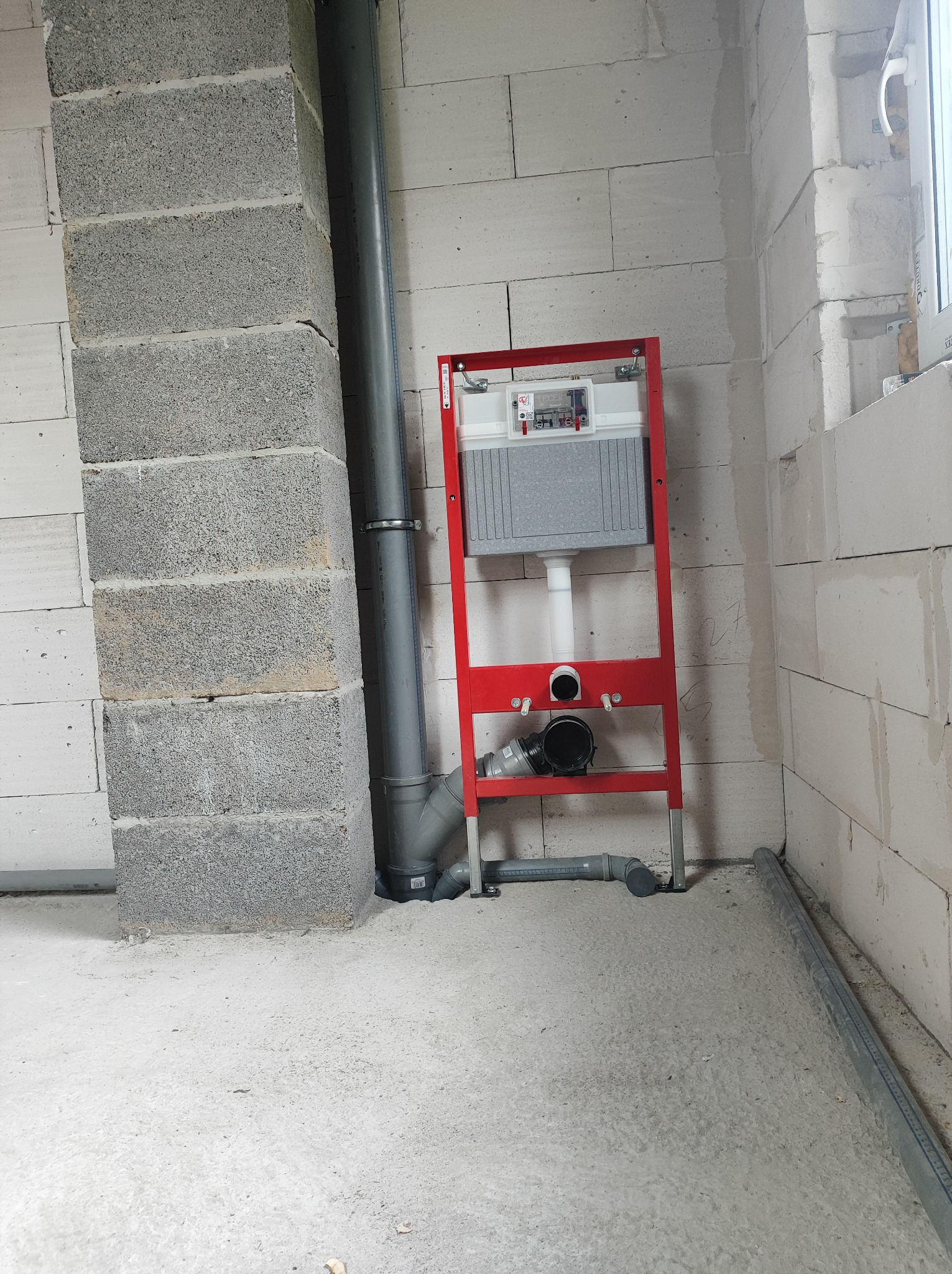 Stelaż podtynkowy WC z widocznymi przyłączami kanalizacyjnymi i rurą odpływową, surowe ściany z betonu komórkowego w tle.