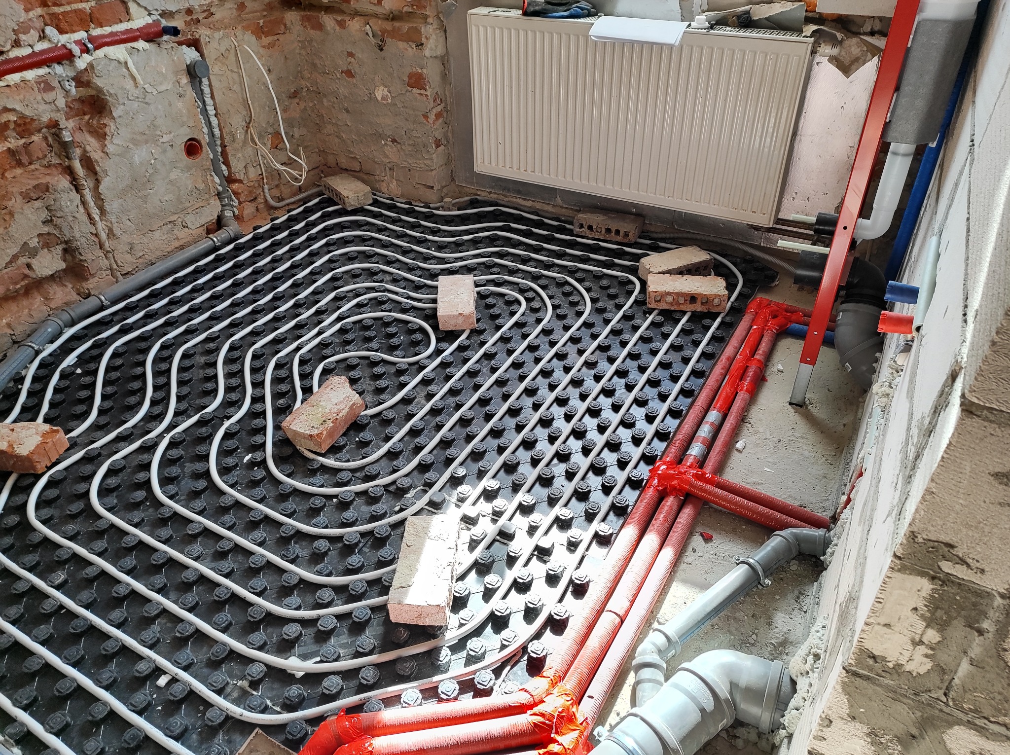 Rozłożona instalacja ogrzewania podłogowego z białych rurek na czarnej macie, przytrzymywana cegłami, widoczne czerwone rury i grzejnik na ścianie w surowym stanie.