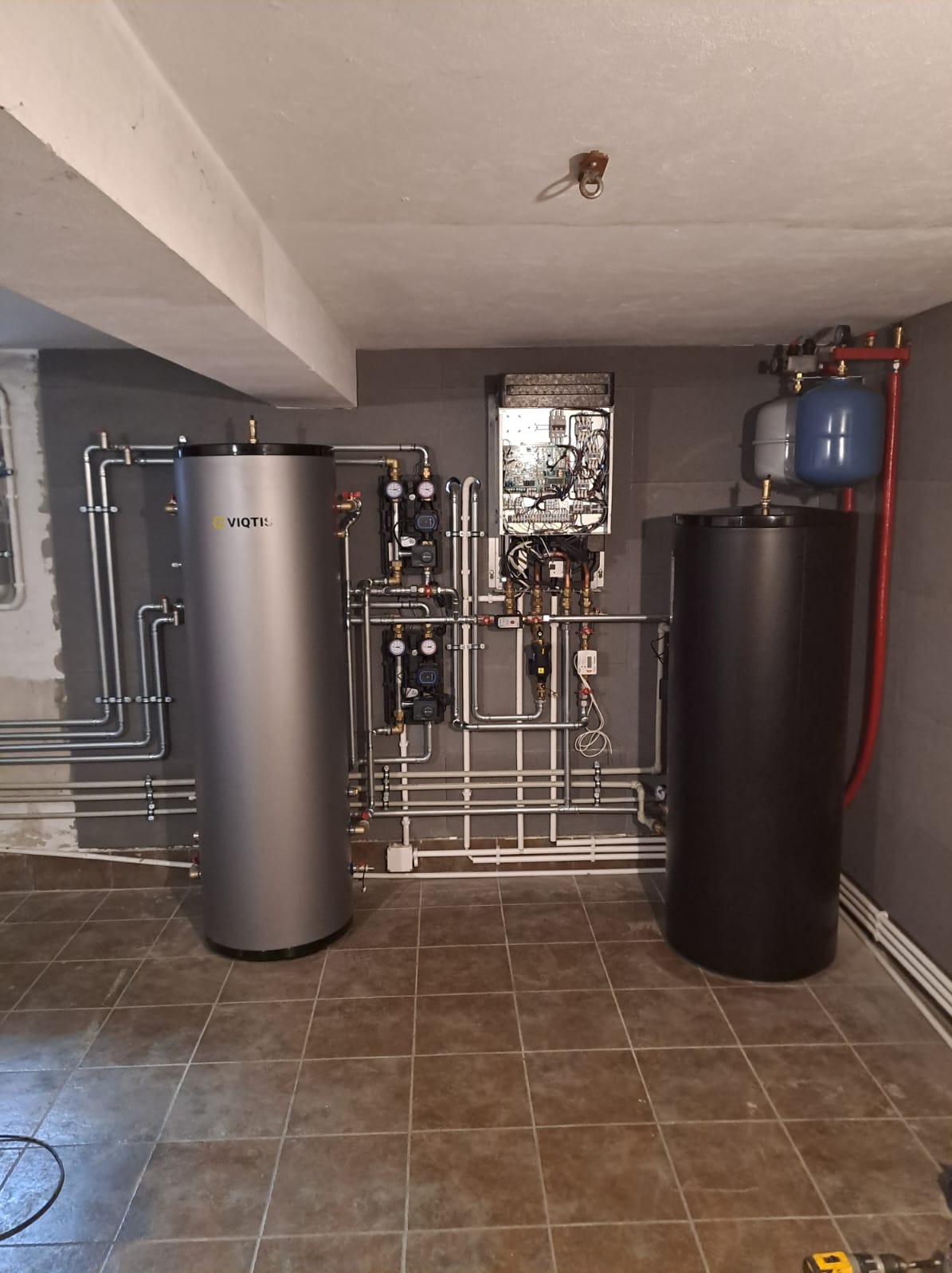 Zmontowana kotłownia z dwoma zbiornikami, srebrnym z logo VIQTIS i czarnym, oraz rozbudowaną instalacją rurową i sterownikiem na szarej ścianie w piwnicy z brązowymi płytkami podłogowymi.