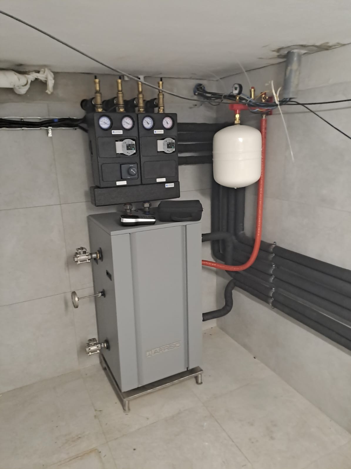 Szary piec grzewczy z czarną armaturą, biały zbiornik wyrównawczy i rury w pomieszczeniu z szarą płytką podłogową i ścienną.