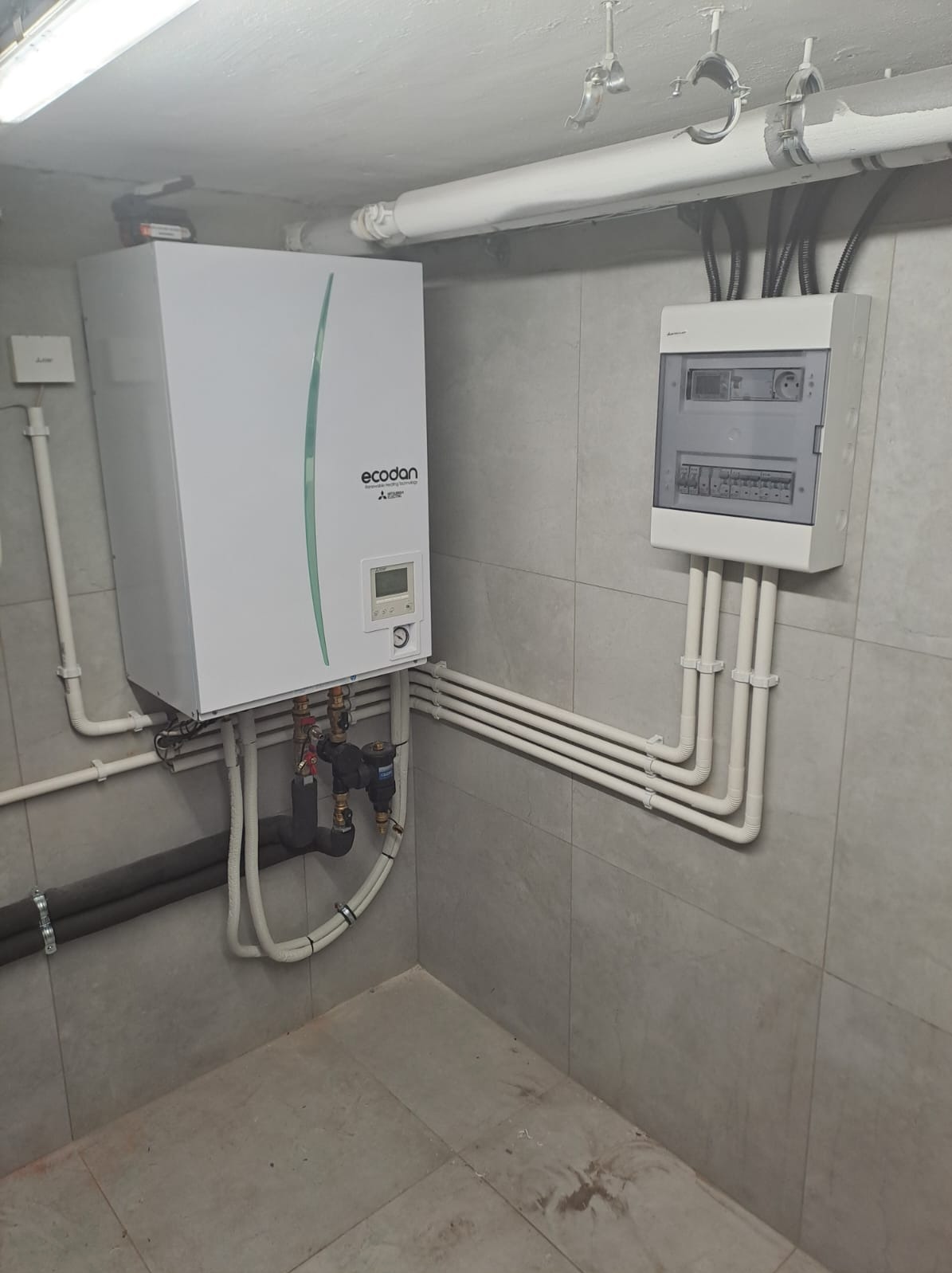 Biała pompa ciepła Ecodan Mitsubishi, zamontowana na szarej ścianie z płytkami, z widocznymi rurami, zaworami i skrzynką elektryczną.