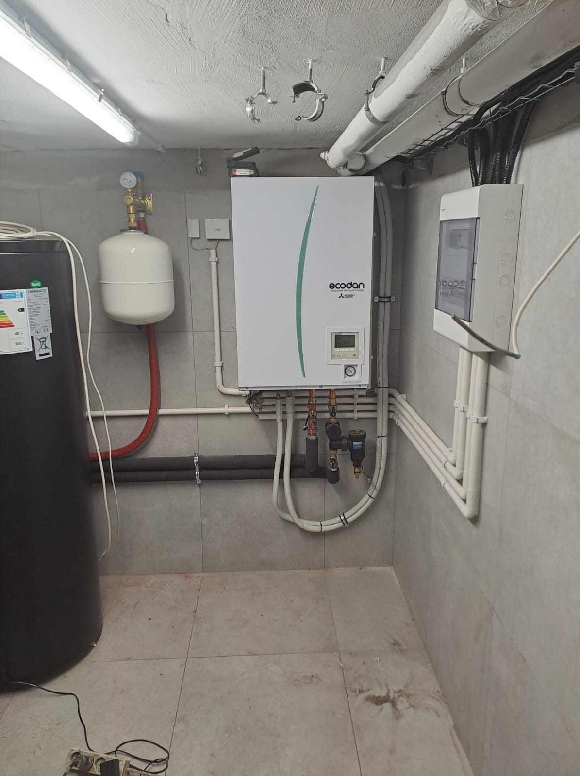 Biała pompa ciepła Ecodan zamontowana na szarej, kafelkowanej ścianie piwnicy, widoczne rury, zbiornik buforowy, skrzynka elektryczna i elementy instalacji.