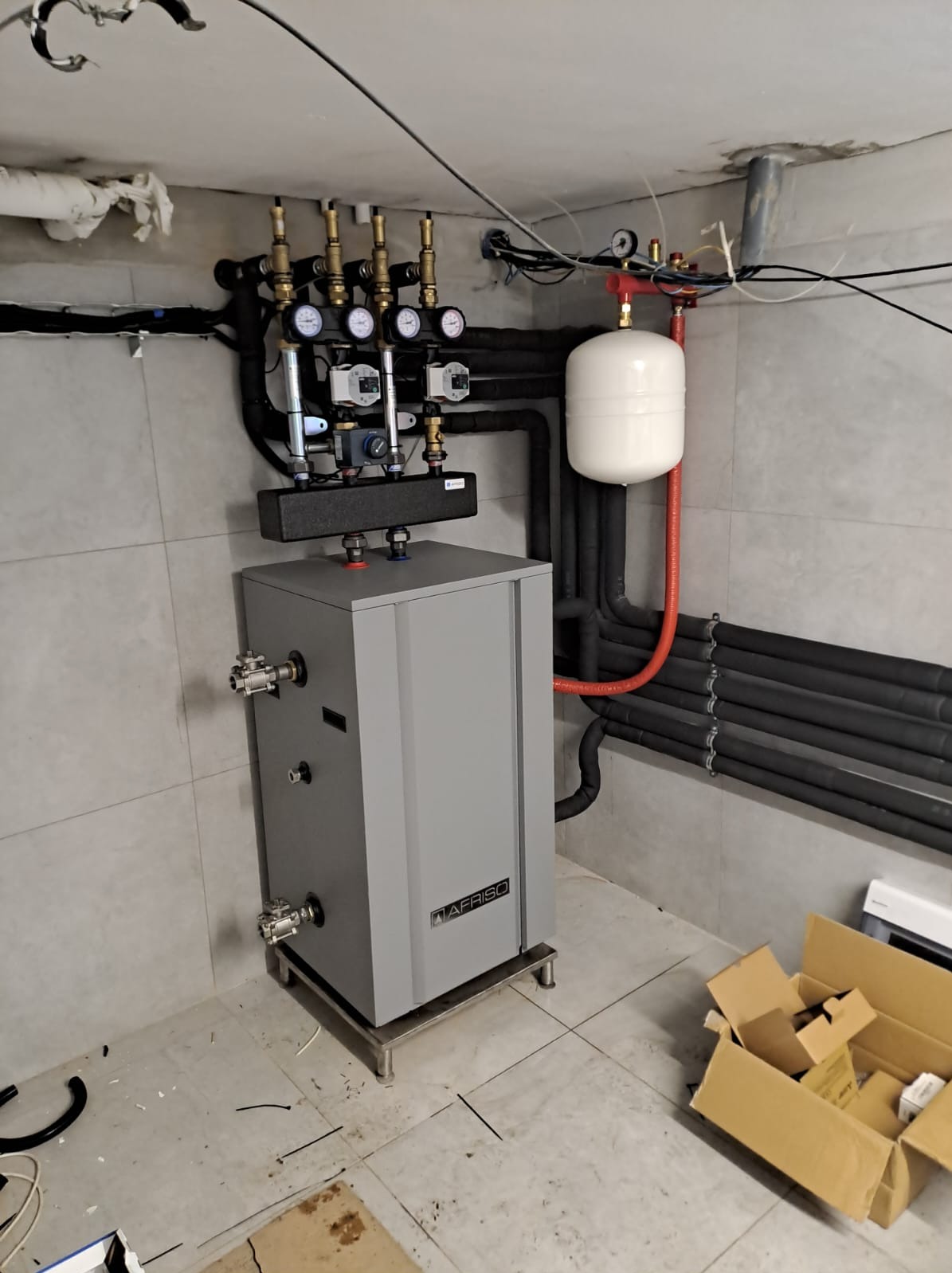 Szara kotłownia marki AFRISO w Krakowie z rozbudowaną instalacją rur, zaworów i manometrów, dodatkowo biały zbiornik wyrównawczy i pozostałości po opakowaniach na podłodze.