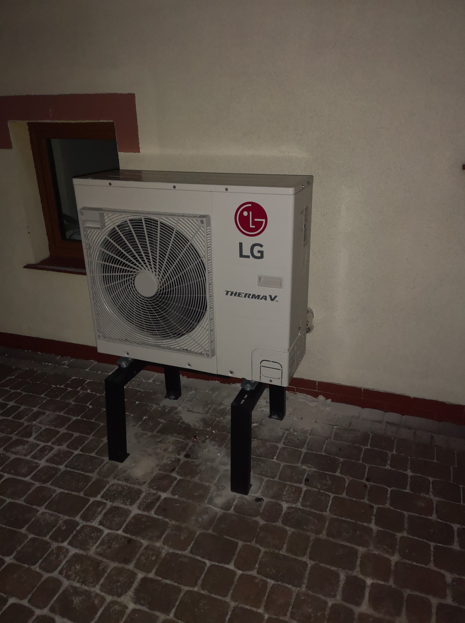 Biała jednostka zewnętrzna pompy ciepła LG Therma V zamontowana na czarnych, metalowych wspornikach na brukowanym podłożu, widoczny fragment okna w tle.