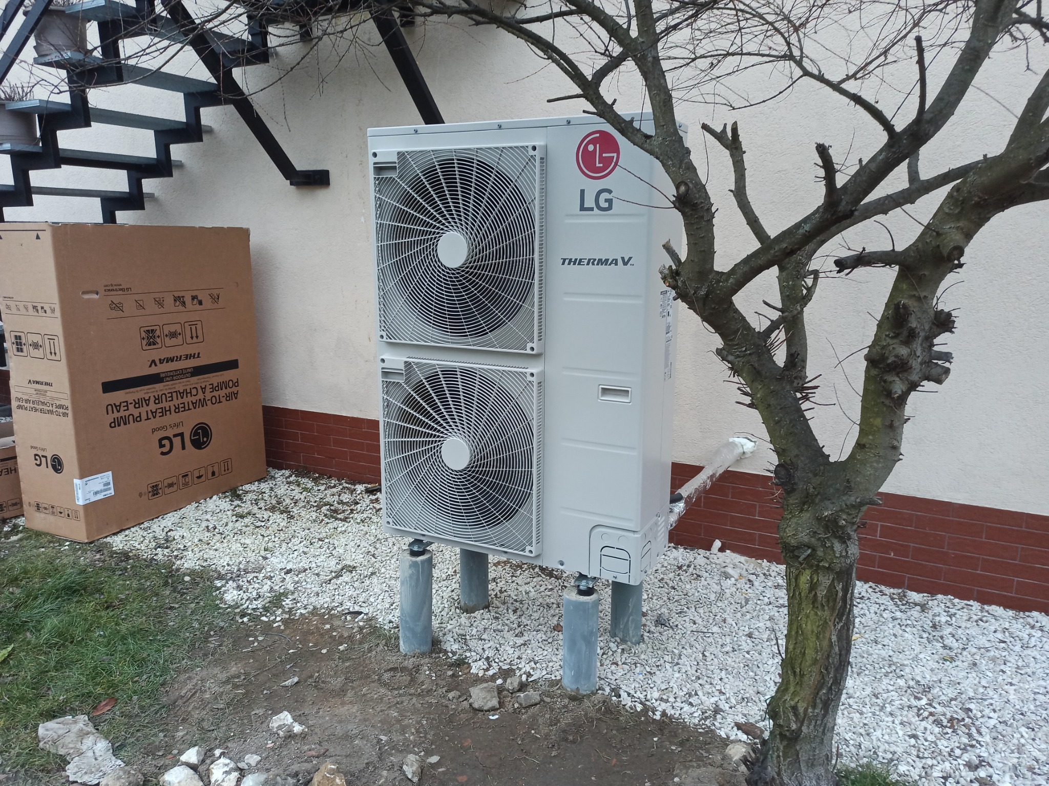 Biała pompa ciepła LG Therma V zamontowana na betonowych wspornikach, obok kartonowe opakowanie, na tle ściany budynku z metalowymi schodami.