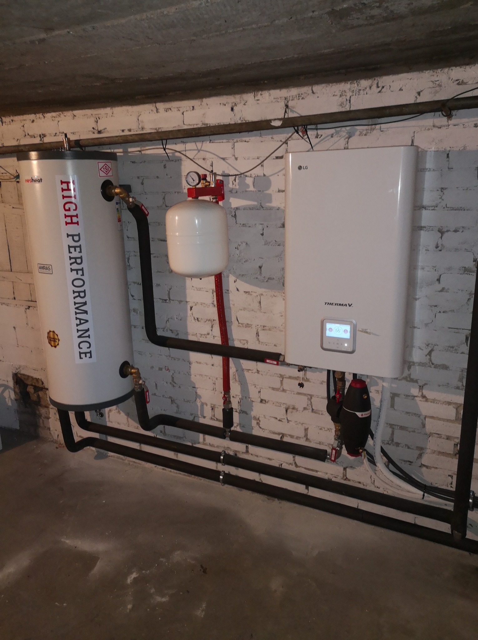 Biały zasobnik ciepłej wody 'High Performance' z czarnymi rurami obok białej pompy ciepła LG Therma V na tle białej ceglanej ściany w piwnicy.