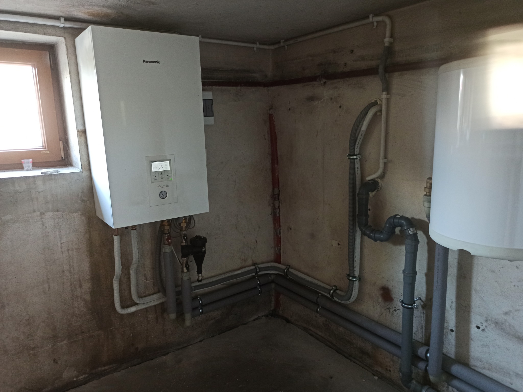 Biała pompa ciepła Panasonic zamontowana na ścianie w piwnicy, widoczne rury instalacyjne i bojler. Ściany z surowego betonu.