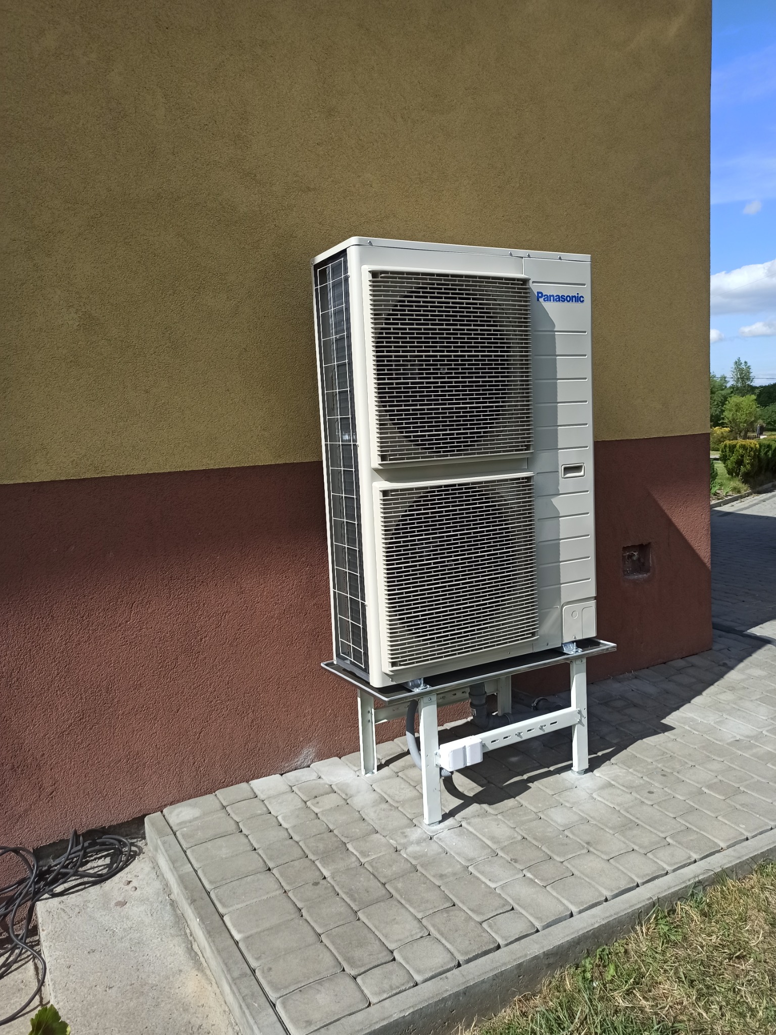 Zewnętrzna jednostka pompy ciepła Panasonic zamontowana na metalowym stelażu przy dwukolorowej elewacji budynku i chodniku z kostki brukowej.
