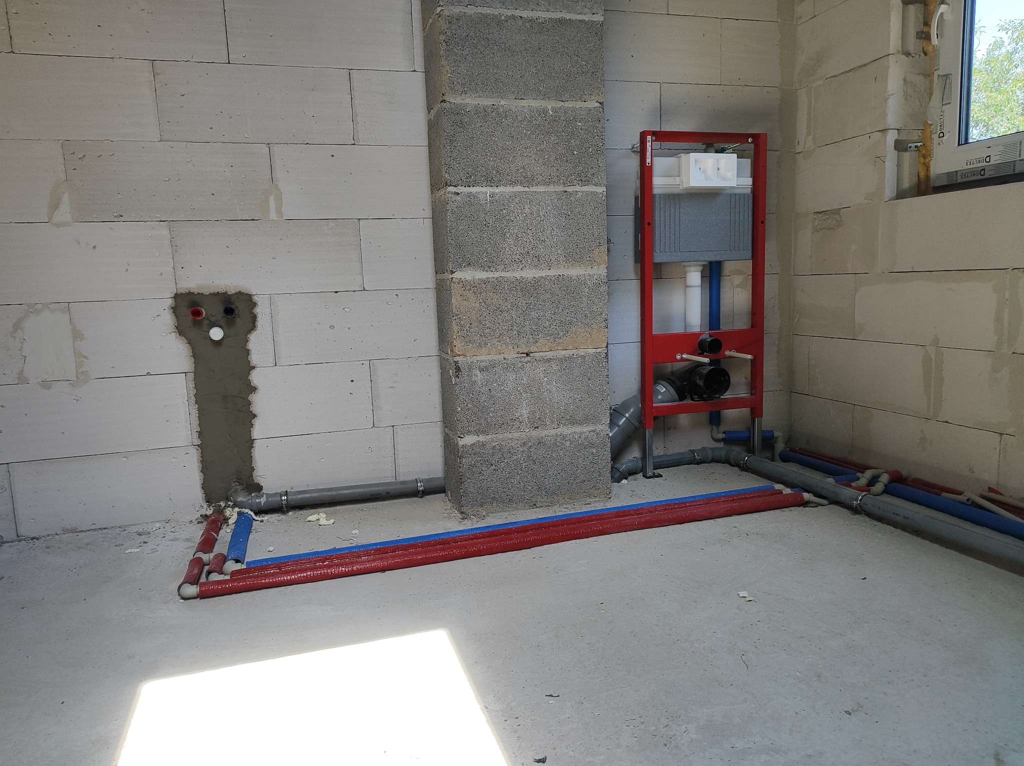 Surowy stan instalacji hydraulicznej w łazience z bloków betonu komórkowego: widoczny stelaż podtynkowy WC na czerwonej ramie, rury doprowadzające wodę w czerwonej i niebieskiej izolacji oraz szare...