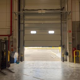 Arkadiusz Ciążyński - Otwarta segmentowa brama przemysłowa z dwoma małymi oknami, widok z wnętrza hali na zewnątrz, w tle fragment placu i barierki.