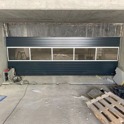 Arkadiusz Ciążyński - Szara segmentowa brama przemysłowa z oknami z siatki, zamontowana w surowym betonowym otworze, widoczny sprzęt i paleta na podłodze.