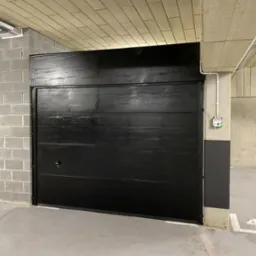Czarna brama garażowa segmentowa, zamknięta, z widocznym zamkiem, w garażu podziemnym z numerem stanowiska 48 na posadzce.