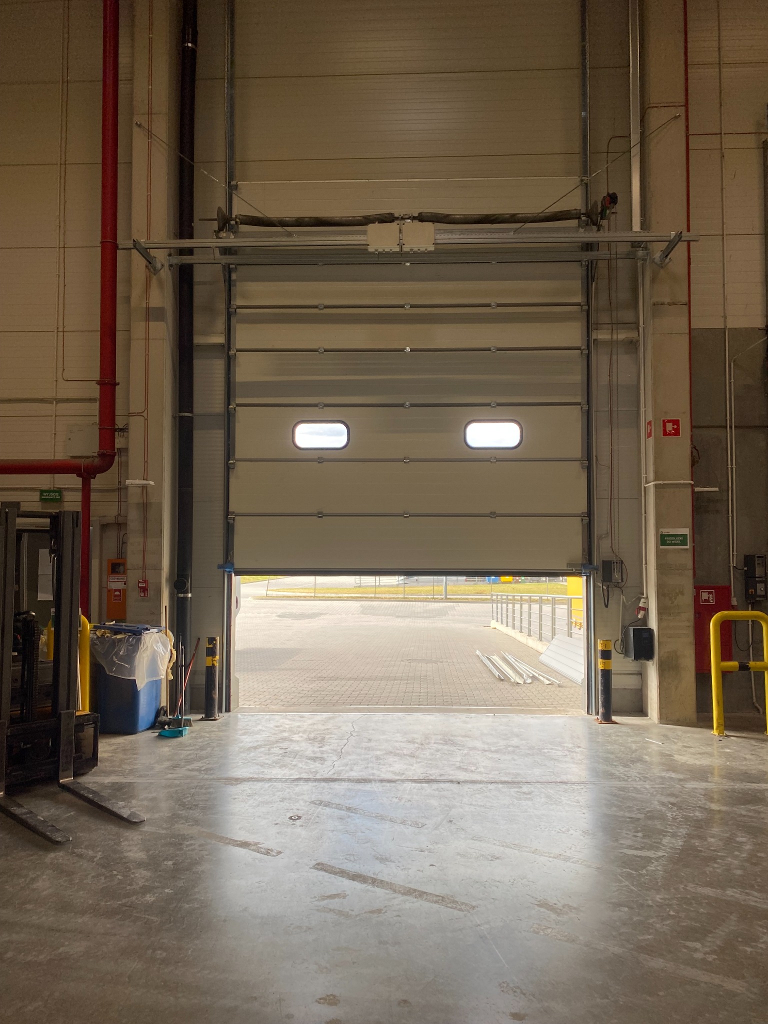 Otwarta segmentowa brama przemysłowa z dwoma małymi oknami, widok z wnętrza hali na zewnątrz, w tle fragment placu i barierki.