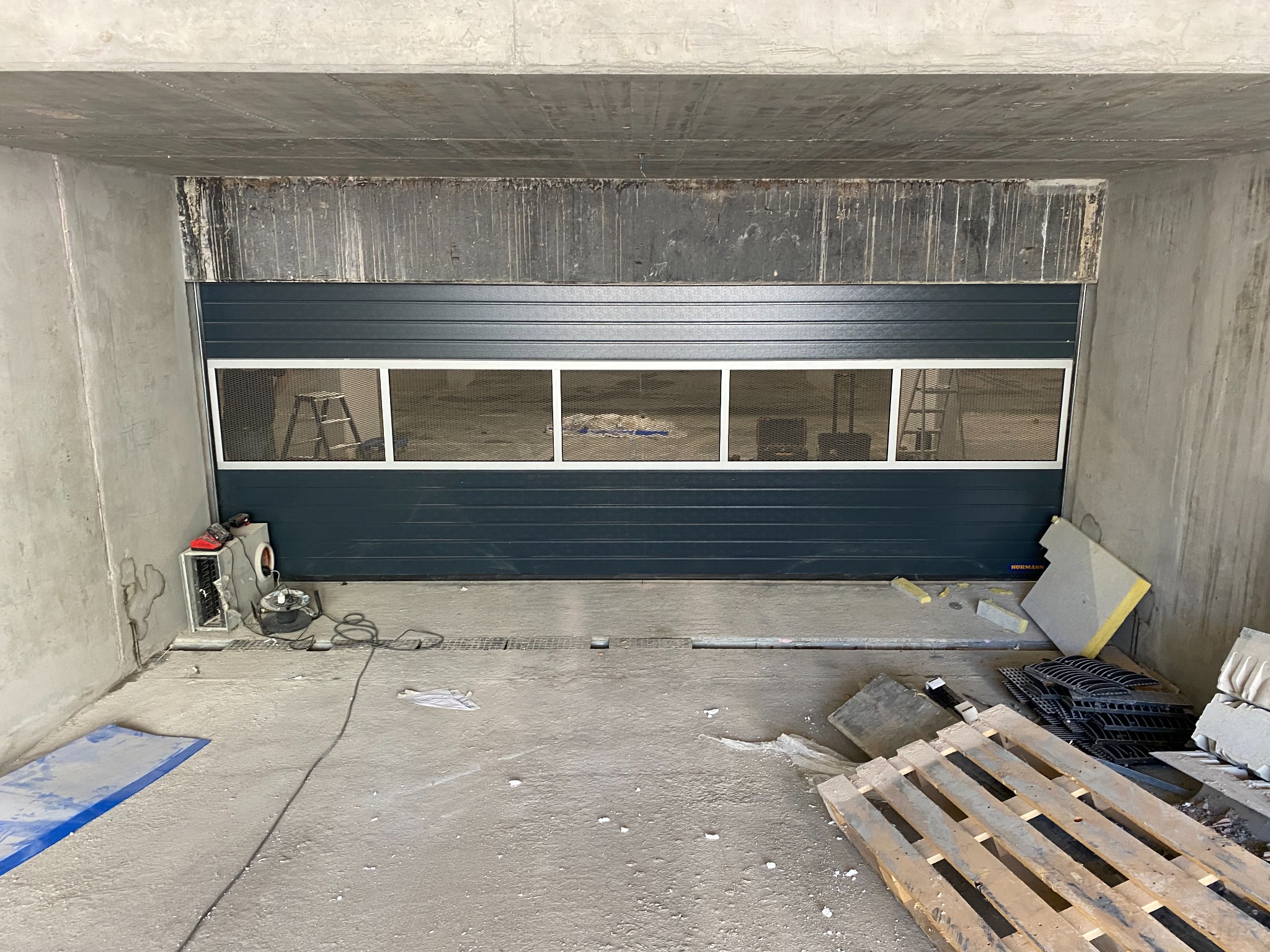 Szara segmentowa brama przemysłowa z oknami z siatki, zamontowana w surowym betonowym otworze, widoczny sprzęt i paleta na podłodze.