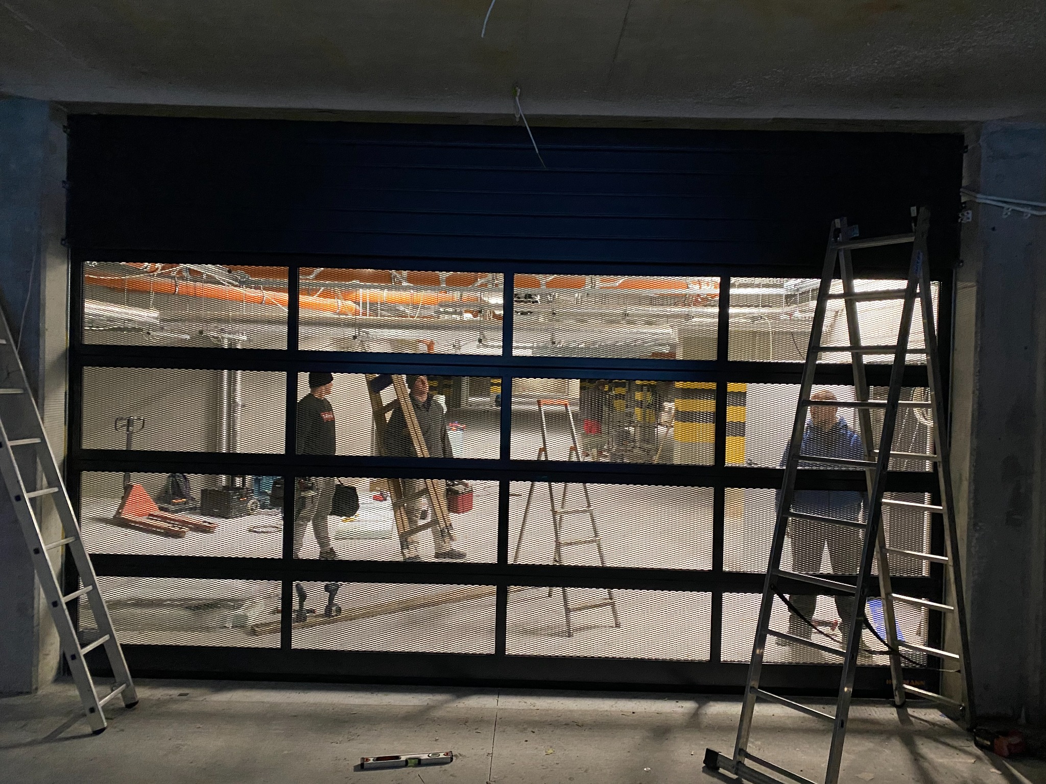 Montaż czarnej, ażurowej bramy przemysłowej z widocznymi pracownikami i drabinkami w surowym wnętrzu hali.