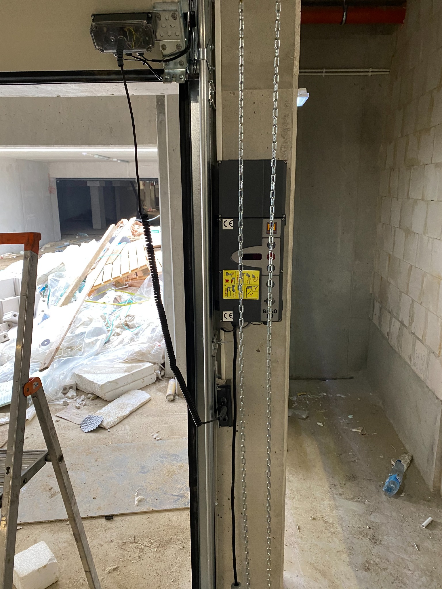 Szczegółowy widok mechanizmu bramy garażowej z łańcuchem, zamontowanego w surowym, betonowym wnętrzu w trakcie budowy, z widoczną drabiną i materiałami budowlanymi w tle.