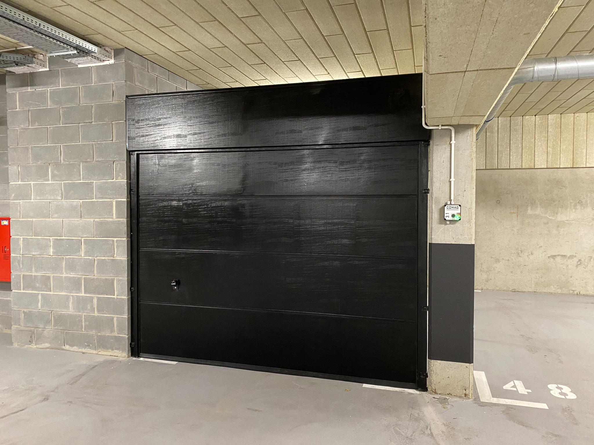 Czarna brama garażowa segmentowa, zamknięta, z widocznym zamkiem, w garażu podziemnym z numerem stanowiska 48 na posadzce.