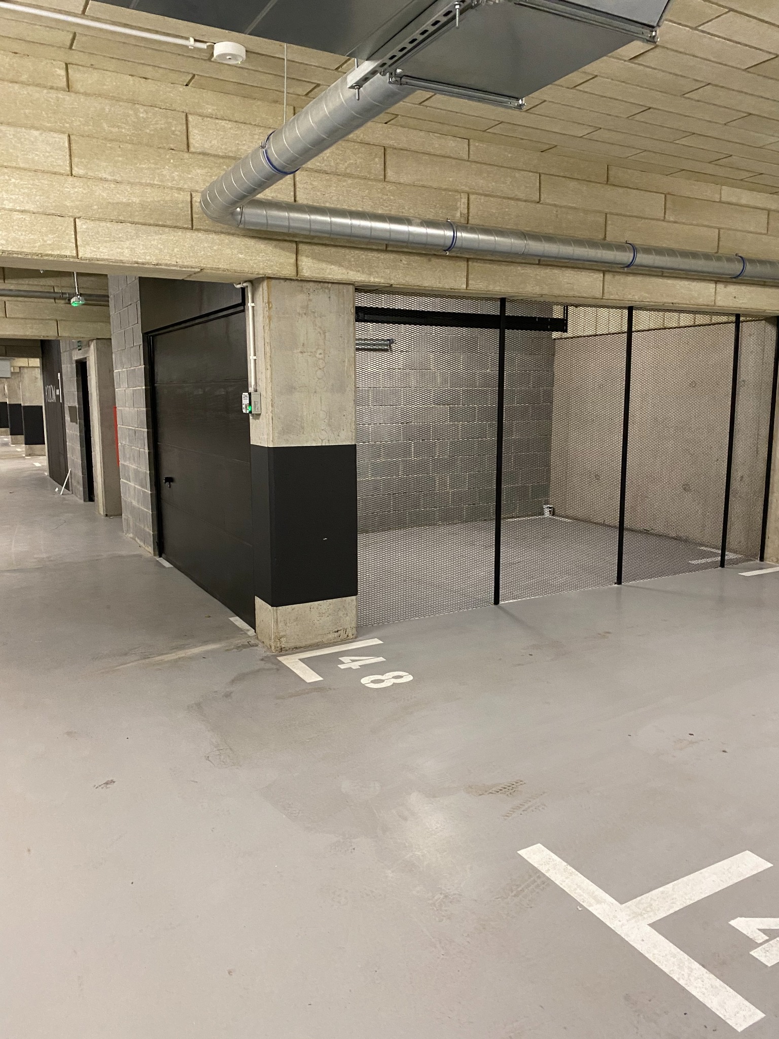 Wnętrze parkingu podziemnego z wydzielonymi miejscami postojowymi za pomocą metalowej siatki i zamykanymi, czarnymi drzwiami.