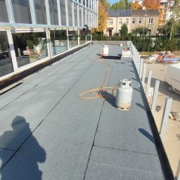 Klaudiusz Rafalski - Płaski dach pokryty szarą papą termozgrzewalną, widoczne butle z gazem i cień osoby, budynek z dużymi oknami w tle.