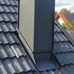 Czarny komin na ciemnoszarym dachu z dachówki ceramicznej, widoczna taśma uszczelniająca, błękitne niebo w tle.