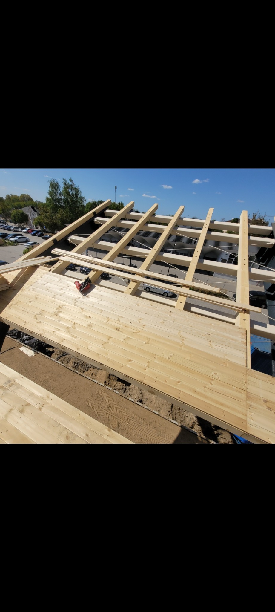 Wznoszenie drewnianej konstrukcji dachu z widoczną więźbą, deskowaniem i narzędziem do montażu na tle błękitnego nieba i zabudowań.