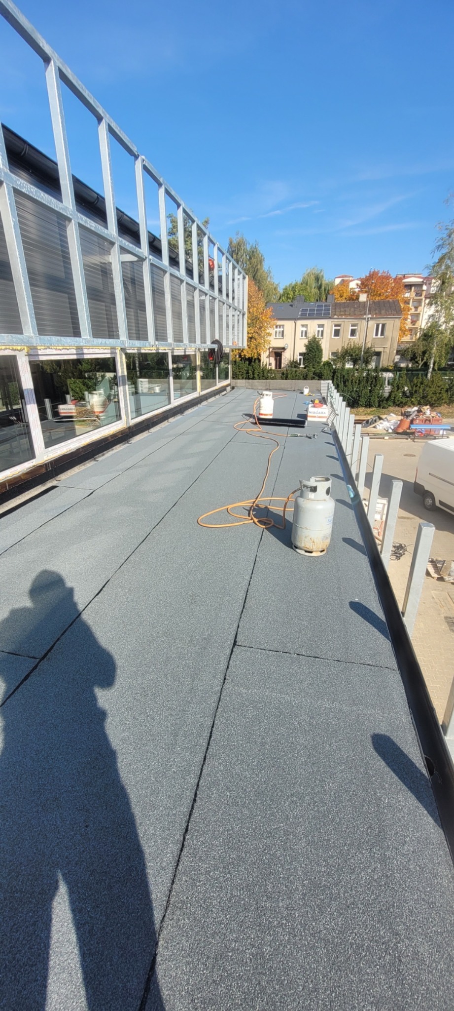 Płaski dach pokryty szarą papą termozgrzewalną, widoczne butle z gazem i cień osoby, budynek z dużymi oknami w tle.
