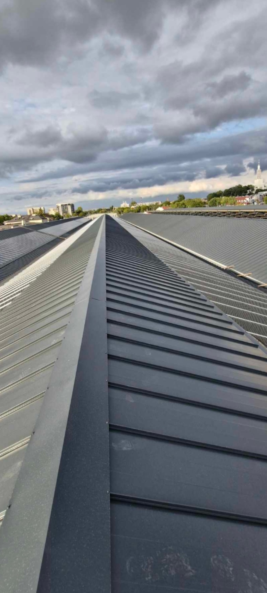 Długi, blaszany dach z wyraźnymi łączeniami, widok z góry na tle pochmurnego nieba i zabudowy miejskiej w oddali.