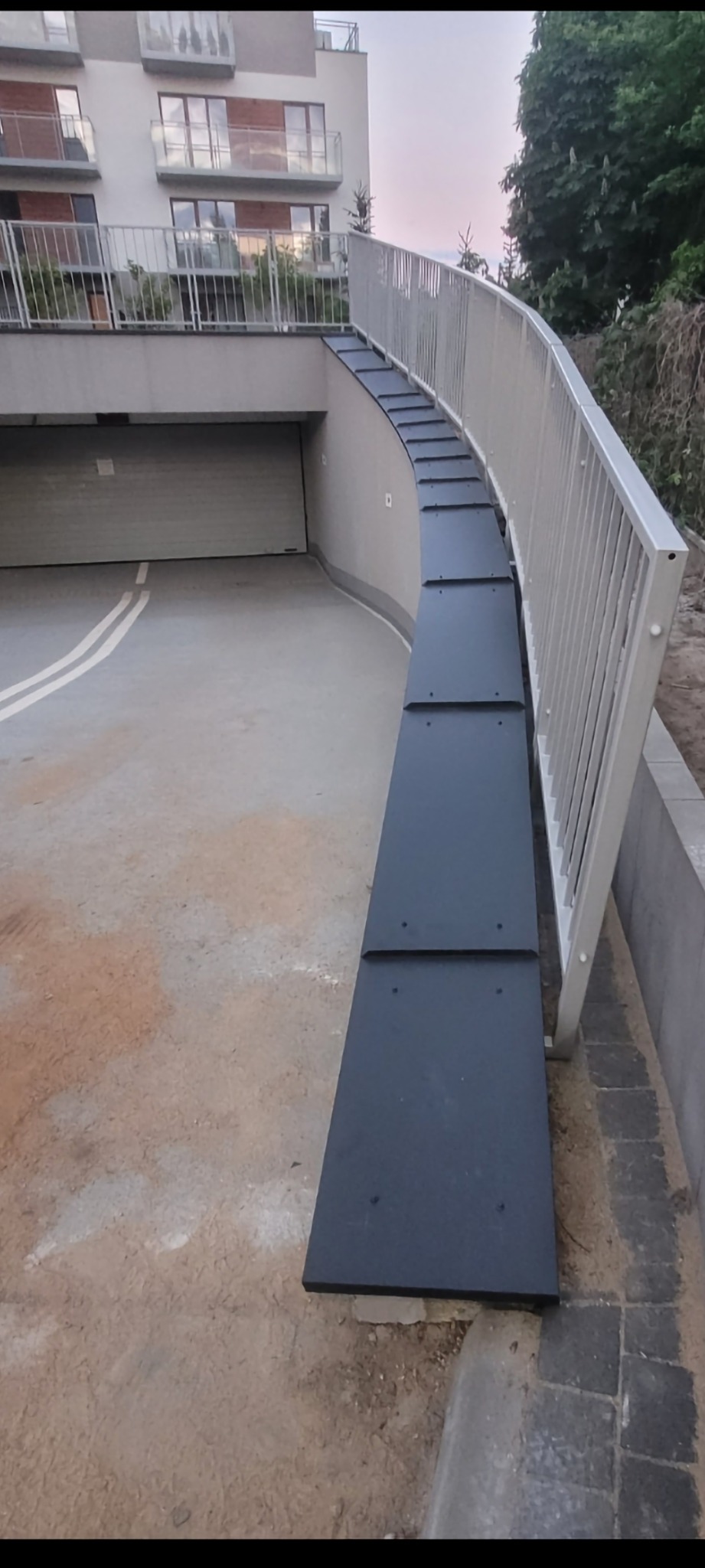 Nowoczesny budynek z rampą dachową prowadzącą do garażu podziemnego, wykończoną czarnymi panelami i białą balustradą.