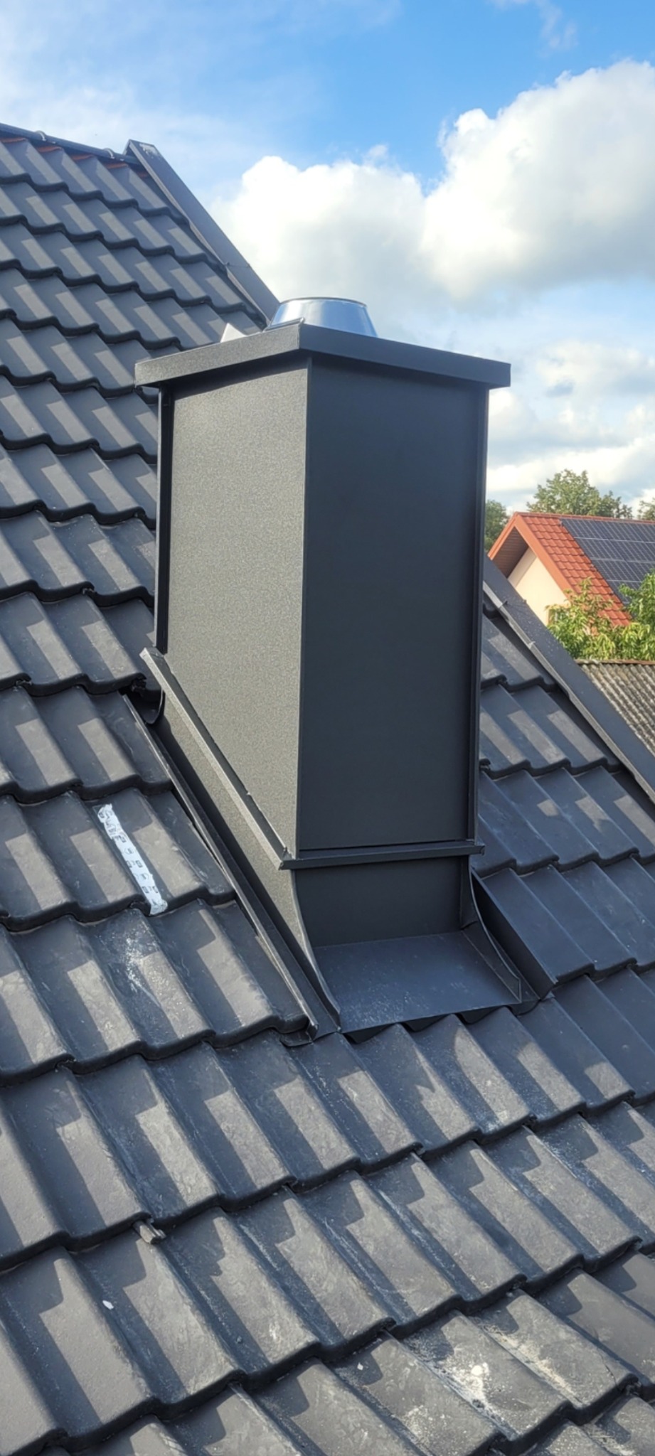 Czarny komin na ciemnoszarym dachu z dachówki ceramicznej, widoczna taśma uszczelniająca, błękitne niebo w tle.