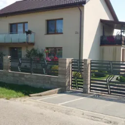 Nowoczesne ogrodzenie posesji z poziomych, szarych profili metalowych na niskim murku z beżowych bloczków, brama wjazdowa z identycznym wypełnieniem, w tle dwupiętrowy dom z balkonem i kwiatami.