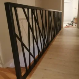 Nowoczesna, czarna poręcz schodowa z geometrycznym wzorem, zamontowana na drewnianych schodach i podłodze; widoczne ślady prac wykończeniowych w tle.