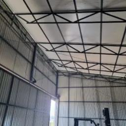 Wnętrze hali z konstrukcją stalową dachu i ścianami z blachy falistej, widoczne okno w oddali.