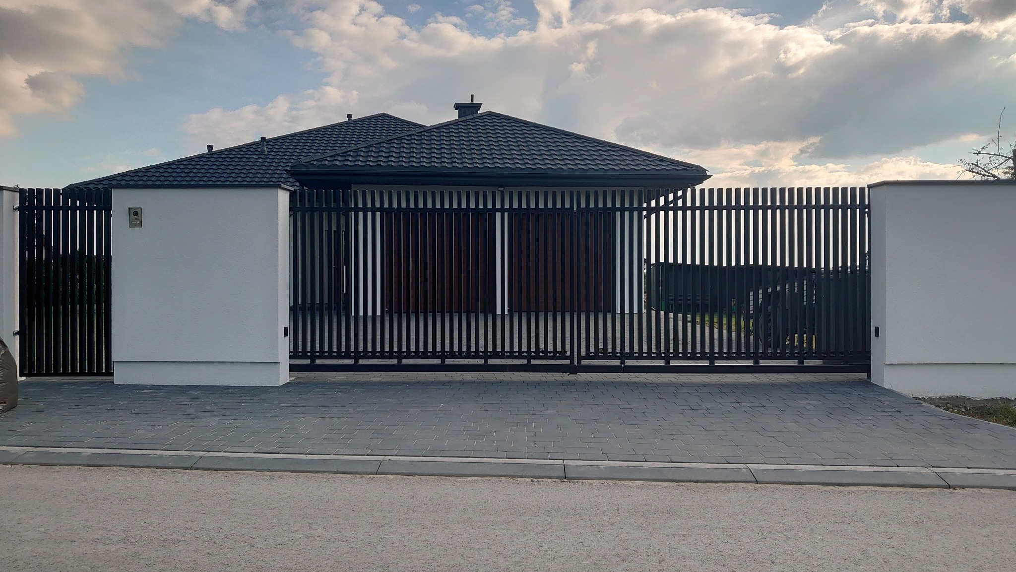 Nowoczesna brama wjazdowa przesuwna z pionowych profili w kolorze czarnym, zintegrowana z białym murem i szarą kostką brukową na podjeździe, widok na dom w tle.