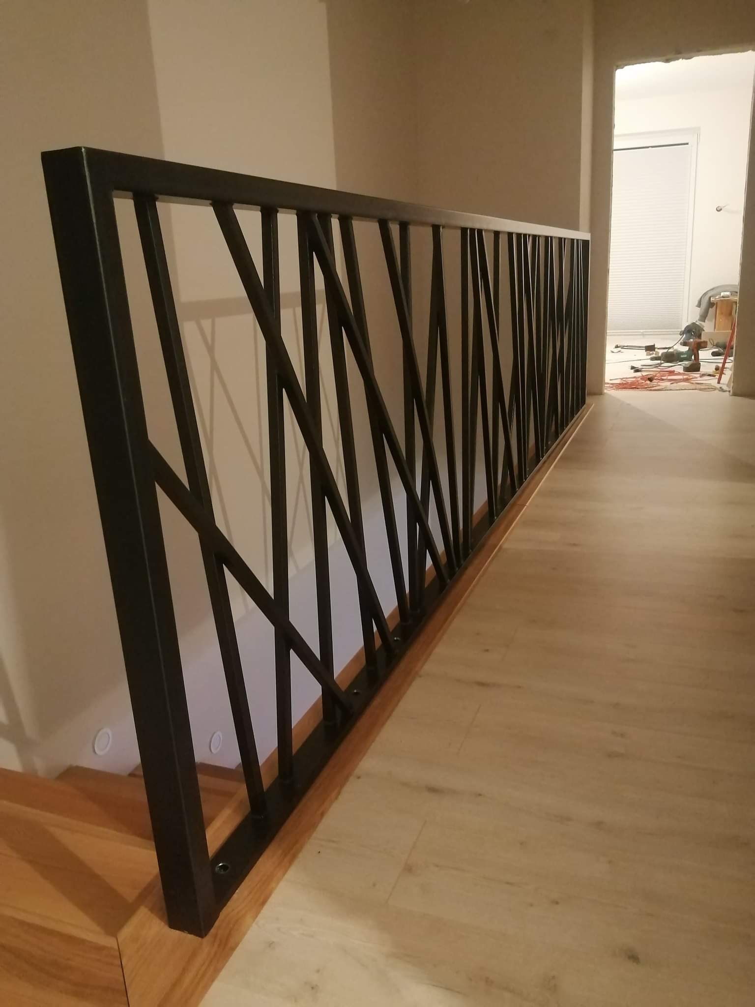 Nowoczesna, czarna poręcz schodowa z geometrycznym wzorem, zamontowana na drewnianych schodach i podłodze; widoczne ślady prac wykończeniowych w tle.