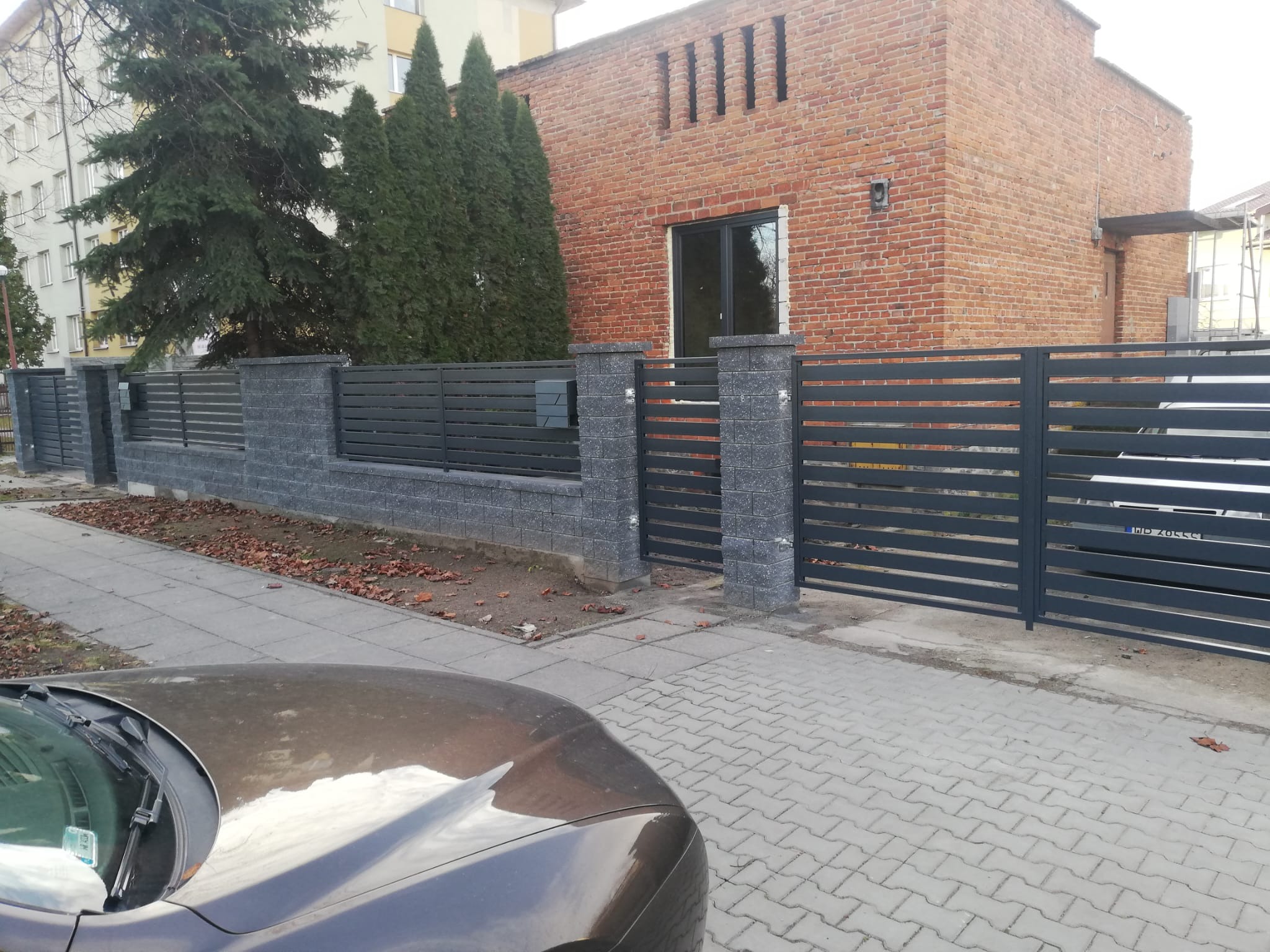 Nowoczesne ogrodzenie z poziomych, szarych paneli i słupków z szarej kostki, przylegające do ceglanego budynku z zielonymi drzewami w tle, widok z ulicy z fragmentem samochodu na pierwszym planie.
